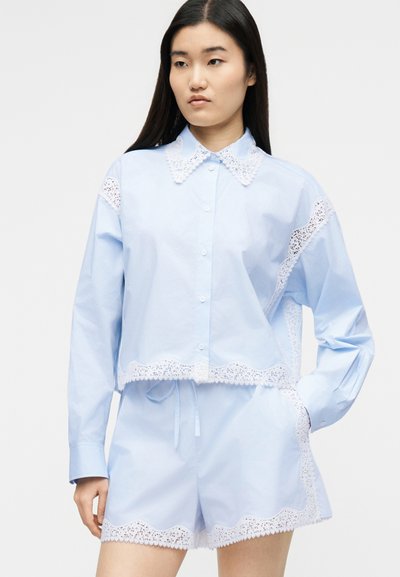 Γυναίκα που φοράει ανοιχτό μπλε πουκάμισο με κουμπιά και ταιριαστό σορτς με λευκή δαντέλα στο γιακά, τα μανίκια και τα τελειώματα, στέκεται με το ένα χέρι στην τσέπη.