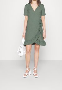 Vihreä kietaisumekko, jossa on lyhyet hihat, röyhelöhelma ja teksturoitu kangas, yhdistetty valkoisiin korollisiin sandaaleihin ja valkoiseen käsilaukkuun.