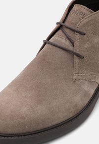 Botín de ante gris con cordones marrones, punta redondeada y superficie texturizada. Los detalles de costura a lo largo de la suela acentúan el diseño.