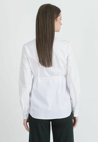 Camicia bianca in cotone con maniche lunghe, dettaglio di legatura in vita sul retro, design strutturato e polsini con bottoni, abbinata a pantaloni scuri.