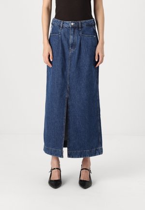 Vrouw die een lange blauwe denimrok draagt met een voorsplit, zwart topje en zwarte puntige Mary Jane schoenen, staand tegen een witte achtergrond.