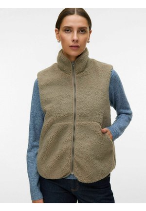 Donna che indossa un gilet in pile sherpa beige con tasche frontali sopra un maglione a maniche lunghe blu e jeans scuri, in piedi davanti a uno sfondo semplice.