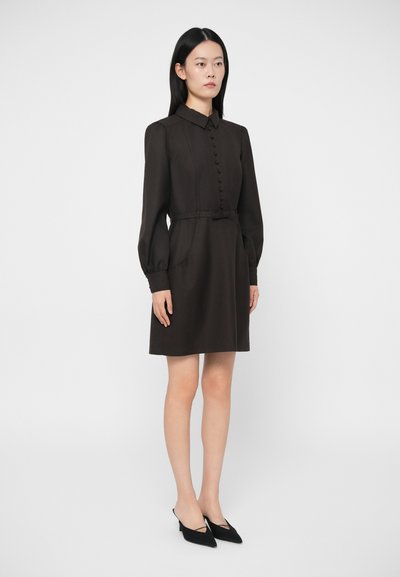 Sieviete stāv cer Melnu kleitu ar garām piedurknēm līdz ceļgaliem, kurai ir pogas un josta, apvienojot to ar melnām zemu papēžu smailām kurpēm.