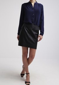 Camicia blu navy con bottoni e maniche lunghe, abbinata a una mini gonna in pelle nera. L'outfit presenta una texture liscia e una silhouette aderente.