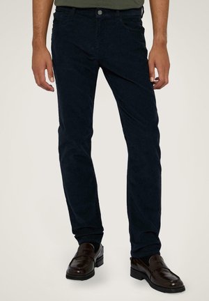 Uomo che indossa pantaloni slim fit blu scuro e mocassini in pelle marrone scuro, in piedi davanti a uno sfondo chiaro.