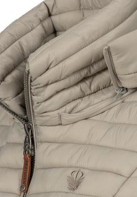 Beige pufferjack met een hoge kraag, voorzien van een doorgestikte textuur, twee ritsen en een decoratief embleem aan de linkerkant vooraan.