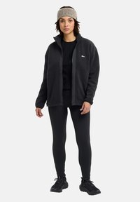 Veste en polaire noire avec deux poches avant, associée à un haut noir et des leggings. Le mannequin porte un bandeau en tricot beige et des chaussures de sport noires.