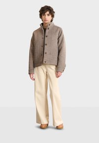 Brown checkered jacket with a high collar, five buttons, and long sleeves, paired with cream wide-leg pants and wooden platform clogs.