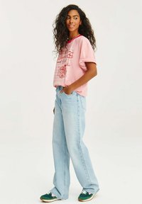 Růžové grafické tričko s ohrnutými rukávy, se červenými vzory, zkombinované s celkově světle modrými širokými džíny a zelenými teniskami s růžovými akcenty.