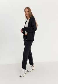 Haut à fermeture éclair noir et joggeurs assortis, en matériau doux. Un crop top blanc en dessous. Modèle portant des chaussures de sport gris clair.