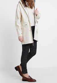 Veste beige légère avec capuche, associée à des leggings noirs à pois et des mocassins marron. Comprend des poches et une coupe décontractée.