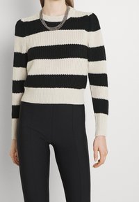 Pull à rayures noir et crème avec une texture côtelée, encolure ronde et manches longues. Porté avec un pantalon noir ajusté et une chaîne en argent à plusieurs couches.