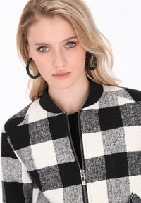 Jacket bomber cu imprimeu în carouri alb-negru, cu fermoar la față, manșete și guler cu textură ribbed. Modelul poartă cercei mari în formă de inel. Detaliu de țesătură vizibil.