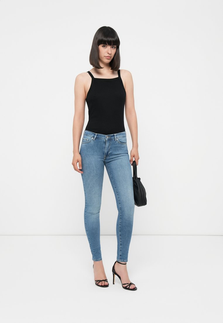 Débardeur noir ajusté, jean slim bleu clair, escarpins noirs à bride. Tenant un petit sac à main noir. Contexte neutre.