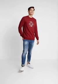 Czerwona bluza crewneck z białym graficznym napisem, zestawiona z jasnoniebieskimi, spranymi dżinsami oraz białymi trampkami. Gładka tkanina, luźny krój.