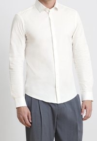 Homme portant une chemise blanche cintrée à manches longues boutonnée et un pantalon gris plissé devant un fond uni.