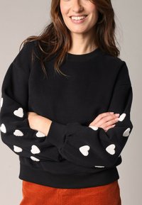 Zwarte sweatshirt met ribgebreide manchetten en zoom, voorzien van witte hartvormige appliqués op de mouwen. Zacht materiaal met een ontspannen pasvorm.