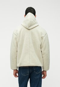 Casaco de fleece cor creme com capuz, apresentando uma superfície texturizada, corte solto e detalhes nas costuras. Usado sobre jeans azuis.