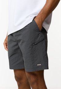 Άνδρας που φοράει σορτς σκούρου γκρι με τσέπη στο πλάι με φερμουάρ και λευκό πουκάμισο, με το ένα χέρι στην τσέπη του σορτς.