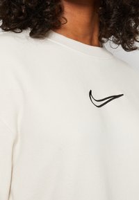 Krämfärgad sweatshirt med ribbad rundhals, prydd med en svart broderad swoosh-logotyp på bröstet, tillverkad av mjukt texturerat tyg.