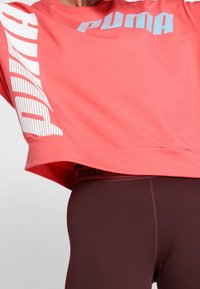 Korálový zkrácený svetr s bílým a světle modrým logem "PUMA" na levém rukávu. Hnědé legíny s páskem s logem.