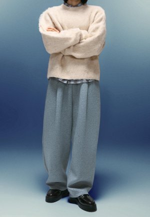 Personne portant un pull pelucheux beige, un pantalon gris à jambes larges, des chaussures noires et une chemise à carreaux partiellement visible au col et à l'ourlet.