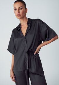 Camicia di raso nera con vestibilità rilassata, maniche corte e chiusura frontale con bottoni. Presenta un colletto e una vita elastica, abbinata a pantaloni coordinati.