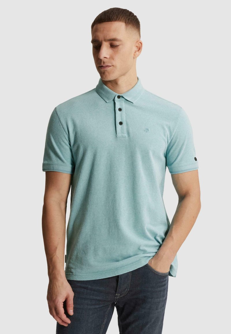 Homme portant un polo bleu clair à manches courtes avec trois boutons noirs et un jean foncé, debout avec une main dans la poche.