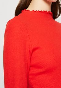 Rotes, geripptes Langarmoberteil mit hohem Kragen und Rüschensaum, das eine glatte Textur und eine figurbetonte Passform aufweist.
