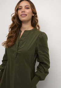 Tmavozelené šaty s dlhým rukávom a mandarin golierom, s gombíkovou légou, riasenými nafúknutými rukávmi a bočnými vreckami. Hladká, ľahká tkanina.