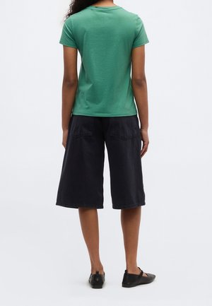Vrouw staat met de rug naar voren gekeerd, draagt een groen kortärmelig shirt, zwarte wijd uitlopende shorts tot op de knie en zwarte platte schoenen.