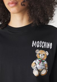 Černé bavlněné tričko s grafickým potiskem medvídka v mašličce s zelenými, červenými a bílými akcenty a textem "MOSCHINO" v bílé barvě.