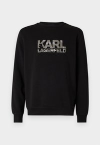 Fekete pulóver, amelyen egy grafikus "KARL LAGERFELD" logó található, félkövér, csíkos ezüst és fehér betűkkel. Klasszikus környakú és bordás mandzsettával.