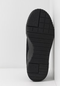 Semelle noire de chaussure avec motifs de prise texturés, incluant des carrés et des lignes horizontales, positionnée contre des murs d'angle blancs.