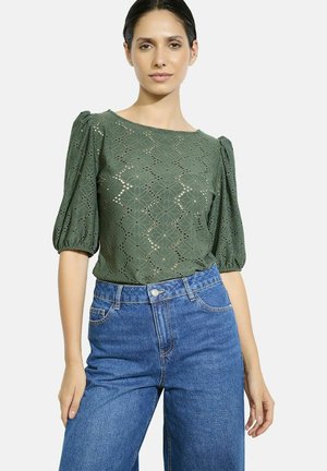 Blusa verde, con trama, dotata di un motivo floreale ritagliato e maniche a sbuffo, abbinata a jeans blu a vita alta.