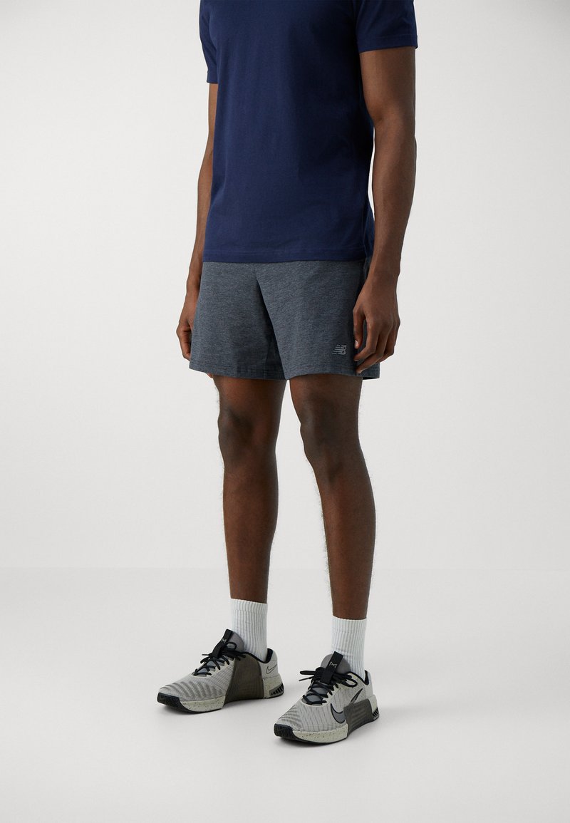 Chemise navy à manches courtes, shorts de sport gris et chaussures à motifs noir et blanc avec des accents noirs. Des chaussettes rayées aux chevilles complètent la tenue.