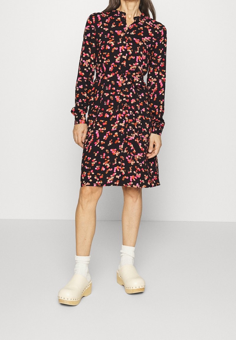 Robe noire avec un motif floral coloré, manches longues, devant à boutons, taille cintrée et ourlet évasé. Associée à des sabots de couleur claire.