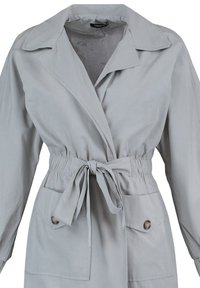 Veste grise avec un col à revers, une taille élastique et une ceinture amovible à nouer. Comprend deux poches avant et un tissu lisse et léger.