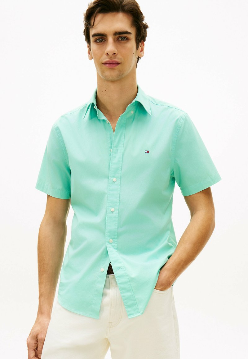 Jeune homme portant une chemise boutonnée à manches courtes vert menthe avec un pantalon blanc, debout avec une main dans la poche sur un fond blanc uni.