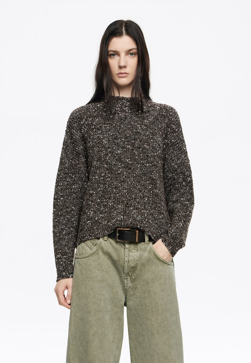 Brauner strukturierter Strickpullover mit hohem Kragen, locker geschnitten mit seitlichen Schlitzen, kombiniert mit lockeren olivgrünen, hoch taillierten Hosen.