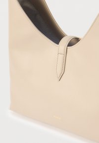 Μπεζ δερμάτινη τσάντα χειρός με στρογγυλεμένες λαβές, εμφανή ραφές και διακριτικό χρυσό λογότυπο «Furla» στο κάτω μέρος μπροστά.