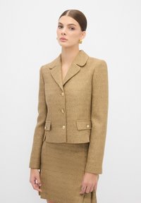 Beige tweed bleiseri, jossa on rakenteellinen kaulus, kultaiset napit ja kaksi taskua, yhdistettynä mukautettuun hameeseen. Pintarakenteinen materiaali koko vaatteessa.