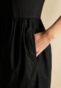 Melnas kleitas ar pieguļošu bodiju un drapētu svārku. Iekļautas sānu kabatas un dekoratīva izšuvuma apmale. Gluda auduma tekstūra.