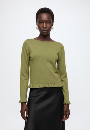 Femme aux cheveux courts portant des boucles d'oreilles en or, un haut à manches longues vert olive avec des bords à volants et une jupe en satin noire.