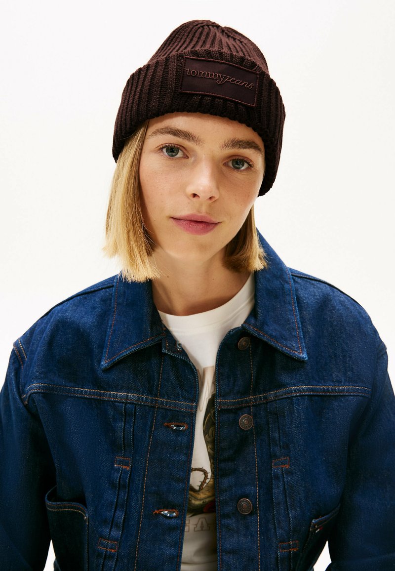 Cappello beanie a coste marrone con un'etichetta logo, indossato da una persona con una giacca di jeans blu, caratterizzata da un classico design con chiusura a bottoni.