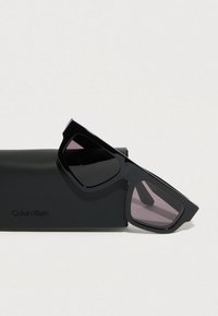 Gafas de sol negras con montura geométrica y lentes degradados, colocadas sobre un estuche negro que lleva el nombre de la marca, Calvin Klein.