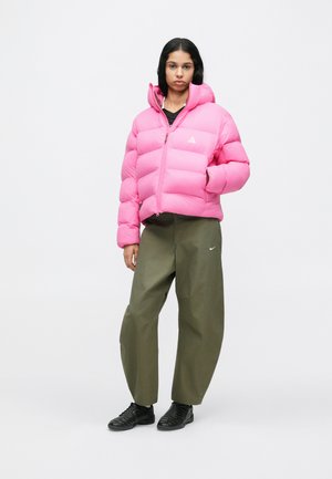 Pink puffer jacket with a high collar, zip closure, and stitched sections. Paired with olive green wide-leg pants and black sneakers.