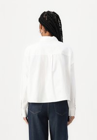 Camicia bianca cropped con maniche lunghe, polsini abbottonati, piega posteriore e colletto, abbinata a jeans in denim scuro.