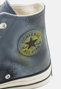 Sneaker alta grigia in canvas con un logo circolare nero e giallo "Converse All Star", occhielli bianchi e una parte superiore testurizzata.