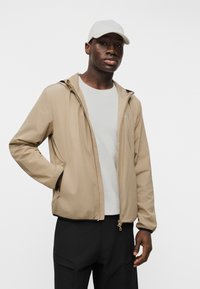 Άνδρας φορούν μπεζ ζακέτα με φερμουάρ και λευκό καπέλο, στέκεται με το ένα χέρι στην τσέπη μπροστά σε λευκό φόντο.